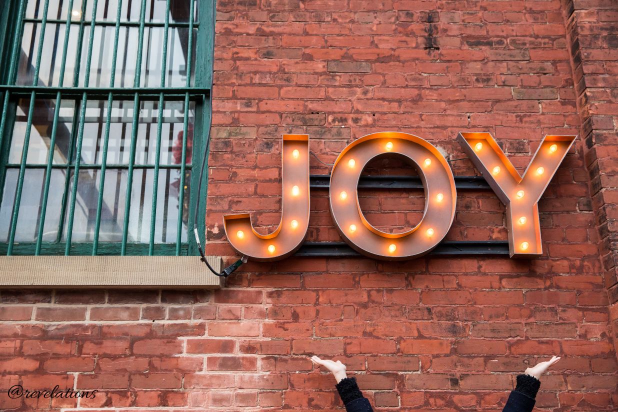 Joy sign light up on brick wall with someone's hands underneath it reaching up